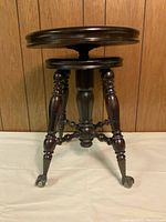 Front angled view of one of the vintage piano stools showing dark mahogany finish, round seat, and ornate carved legs with caster wheels.