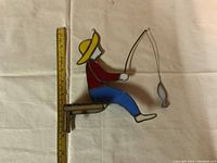 Handmade stained glass fisherman sitting on a wooden dock holding a fishing rod with a fish, shown with measuring tape for size reference