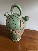 Green ceramic jug with a large loop handle, two spouts, and textured relief decoration with black outlining on the body.