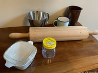 Shows a large wooden rolling pin, stainless steel bowl, enamel mug, brown pitcher, small glass jar with yellow lid, and CorningWare dish with lid on wooden surface.