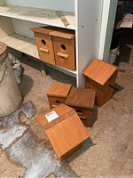 Six wooden bird houses of different simple designs grouped on concrete floor and shelf in basement area