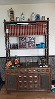 Front view of mid-century modern wooden cabinet with three shelves, holding glassware, steins, and books. Lower storage with multiple drawers and cabinet doors.
