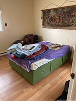 Green captain's bed with six drawers, bedding and blankets on top, in a beige walled room with wooden flooring and tapestry on the wall