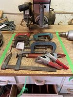 Clamps arranged on a wooden workbench underneath a radial saw, showing assortment and relative sizes