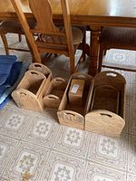 Photo showing seven wooden crates of various sizes with cutout handles, arranged on a patterned vinyl floor near a wooden table and chairs.