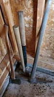 Tools leaning against shed wall on wooden floor: pickaxe, axe, and two sledge hammers