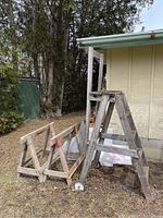 Photo showing two wooden saw horses and one wooden ladder/stand set outside by a building. Saw horses have triangular support frames with metal brackets and faded paint. Ladder has two wheels attached at base for mobility.