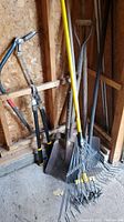 Photo of rakes, shovels, and garden weasel in corner of shed showing handles and metal working parts.