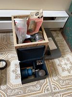 Photo showing a cardboard box with small metal parts, a blue welding tool, and a grey container on a patterned surface.