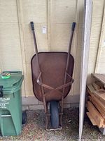 Full view of rusty metal wheelbarrow with tubular handles and single front tire standing vertically against wall.