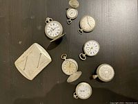 All six antique pocket watches and one cigarette case arranged on dark surface to show overall condition and quantity.