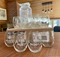 Photo of the clear glass beverage dispenser jar and 16 stemless wine glasses stacked on cardboard with wood panel background, showing embossed 'Nobilo' glasses in front and dispenser behind.