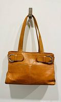 Front view of camel leather tote bag hanging from wall hook, showing two slip front pockets with silver-tone buckle details and double shoulder straps.