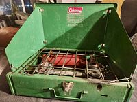 Open view of green Coleman camp stove showing two metal burners, protective grate, and hinged lid with Coleman instructions inside.