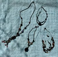 Overview of four costume jewellery pieces on a light blue textured fabric background, including two necklaces and two bracelets all in black tones with brown and red bead accents.