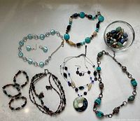 Wide view showing multiple necklaces, a pair of earrings, several bracelets, and a bowl of loose blue and green beads.