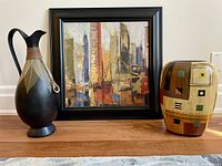 Set of three decorative accessories including a framed abstract painting, ceramic vase, and metal pitcher on wooden floor.