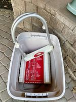 Photo of the folded emergency escape ladder stored in a plastic container with visible metal hooks and instruction label.