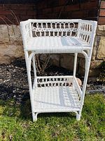 Front view showing the 2 tier wicker shelf, woven loop pattern around top tier, white color, standing on grass outside.