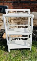 Photo of the front view of the white wicker rattan three-tier shelf unit placed outside on grass with a brick and stone wall background.