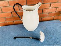 White enamelware jug with black handle and matching ladle with black handle set on blue surface in front of brick wall