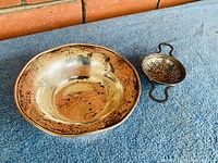 Sterling silver bowl and tea strainer placed on blue fabric showing tarnished surfaces and overall shape.