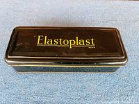 Top view of closed rectangular tin first aid kit with Elastoplast branding in yellow on brown background.