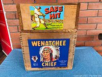 Front view of the two wooden crates stacked with their paper advertising labels visible, showing colorful vintage graphics.