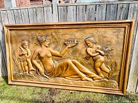 Full view of the large bronze bas relief decoration against a wooden fence on grass, showing overall dimensions and framed surroundings.