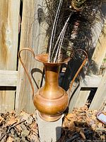 Hammered copper floor vase with double handles filled with decorative peacock feathers against a wooden fence background.
