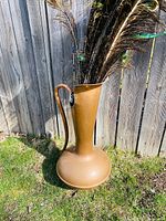 Full view of the large copper jug standing outside on grass with peacock feathers inside, showing the jug's shape and size.