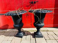 Front view of two black painted cast iron urn planters with classical motif on flared rims on paving stones