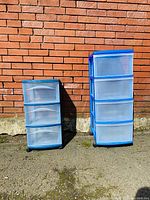 Front view of two plastic storage units, one with 3 drawers and one with 4 drawers, both blue framed with translucent drawers against a brick wall.