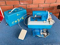 Vintage blue Sister electric sewing machine with foot pedal shown alongside matching blue hard carrying case and instruction manual on blue carpeted surface, brick wall background.