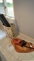 Photo showing Toro 700 Rake-O-Vac blower in box with collection bag, orange extension cords, and instruction manual on tiled floor by window.