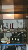 Shelf showing a variety of candle holders including pewter, red glass, clear crystal, Lenox bowl, and brass holders.