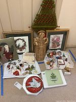 Photo showing assorted Christmas-themed home decor items including framed art prints, fabric ornaments, macrame wall hanging, collector plate, and small trinkets.