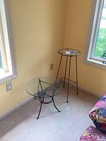 Two glass top tables positioned near a wall and windows, showing different metal frame base designs and glass tops.