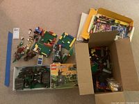 Multiple Lego minifigures laid out on carpet surrounding a partially assembled Lego set on green baseplates and two Lego boxes containing loose parts and instruction booklets.