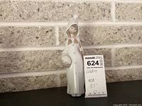 Full view of Lladro figurine standing against a brick wall with price and measurement tag visible, showing the entire figure and base.