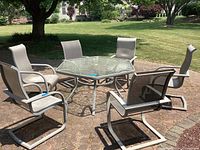 Outdoor photo showing octagonal glass top patio table with metal frame surrounded by six matching chairs with metal frames and mesh fabric seats and backs on a stone patio.