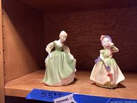 Two Royal Doulton figurines placed on a wooden shelf, Babie on the left in a green dress and Fair Maiden on the right in a pale dress with bonnet.