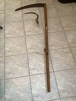 Full view of scythe and sickle laid on a tile floor showing wooden handles and metal blades with visible rust, scythe handle is loose at a joint
