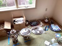 Wide shot showing most of the silver plate items including coffee urn, serving trays, boxes with packing materials, and various pieces laid on the floor next to a window.