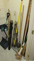 Photo showing Scotts green metal broadcast spreader with two black wheels and some rust, alongside several garden tools including wooden-handle rakes, shovels, a broom, mop, and rolled garden netting.