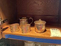 Four-piece set with two ornate napkin rings and two simpler baby cups placed on wooden shelf, viewed from distance.