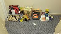 Wide shot showing various boxed and loose tools, bulbs, clamps, and packing supplies arranged on carpeted floor.