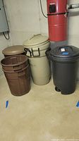 Six plastic trash cans in brown, beige, and black colors; three have lids and handles, three do not; visible wear and soiling.