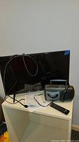 Front view showing LG 27-inch TV, remote control, user manual, power cord, and Venturer portable radio cassette recorder on white shelving unit.