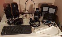 Photo showing assortment of electronics including speakers, cordless phones, stereo system, keyboard, headphones, and power bar on a table.